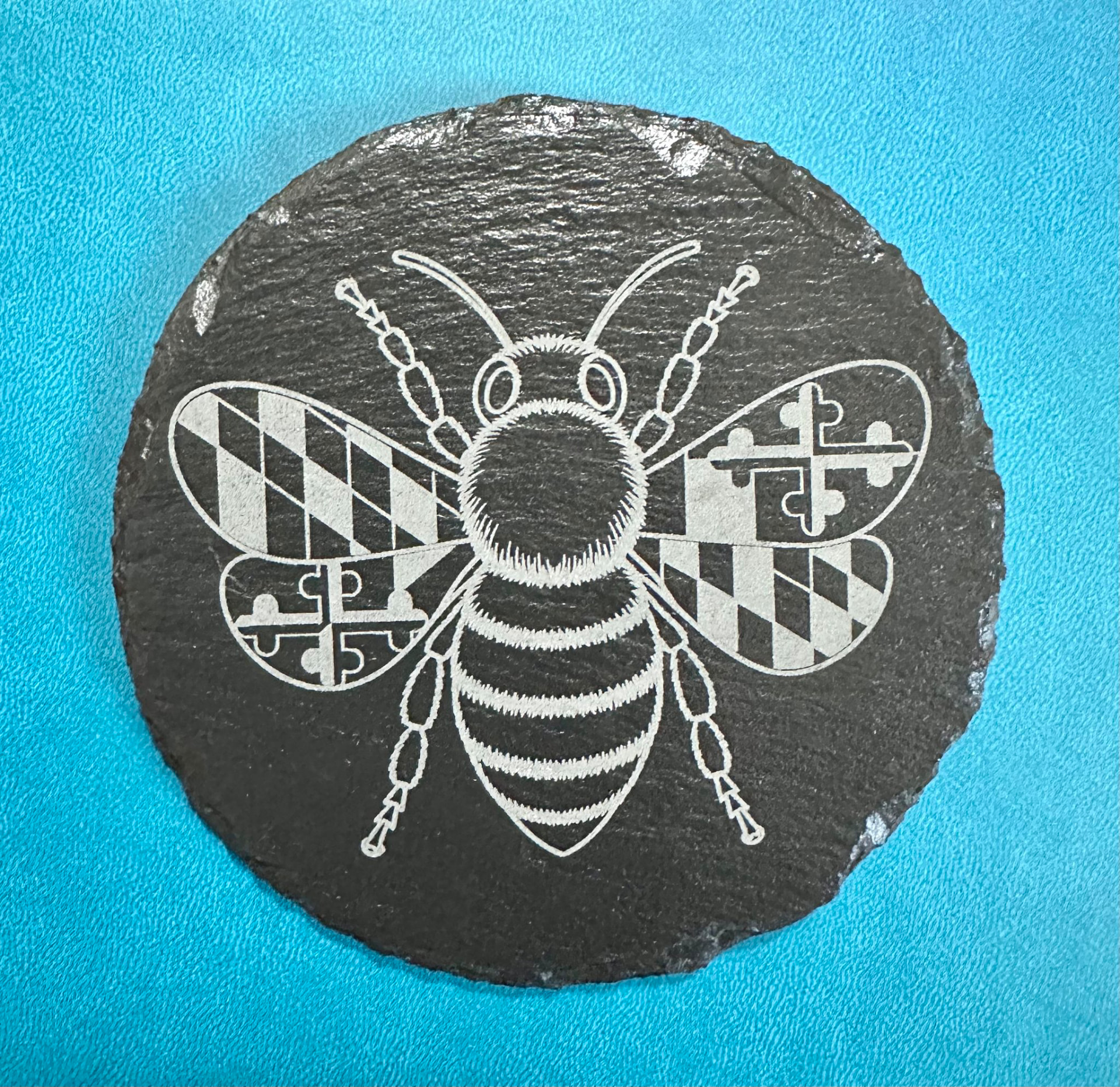Round slate coaster engraved with a bee whose wings carry the Maryland flag